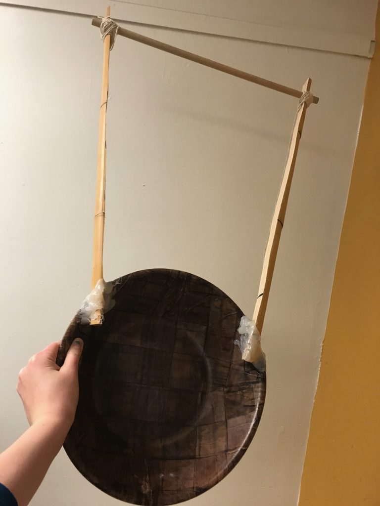 Assembled Frame of Second Prototype, a dark wooden bowl held upright, attached to it  with hot glue are two bamboo rods sticking up like bunny ears, these are the lyre arms, the crossbar is also bamboo and suspended between them, attached to the arms with twine