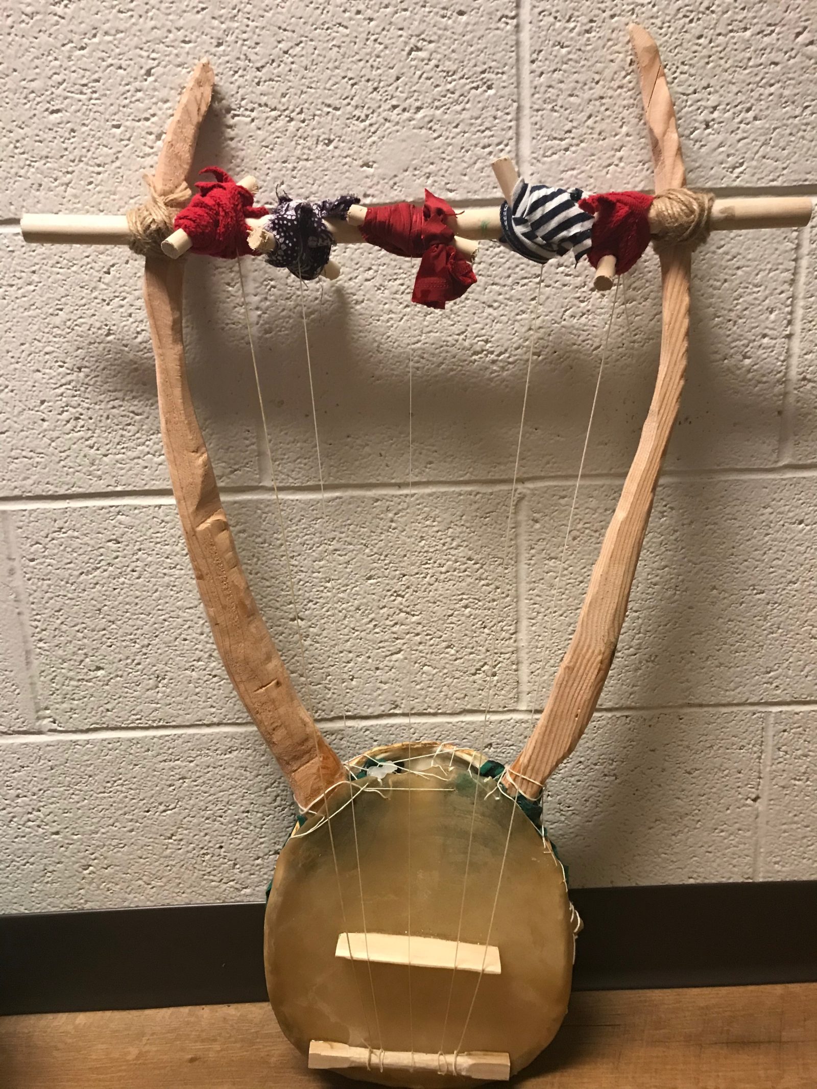 Image of my final lyre, made of goat skin and turtle skin drum, with two carved lyre arms and crossbar, 7 sheep gut strings attached to crossbar with round pegs and old recycled fabric. Only 5 strings are attached as I have to re-string them